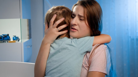 Portrait of young loving mother soothing and caressing scared little boy in children room at night....
