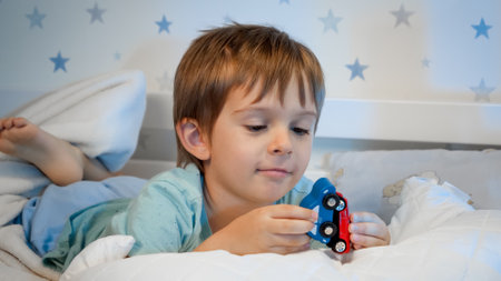 Portrait of little smiling toddler boy lying in bed at night and playing with two wooden toy cars before going to sleep.の写真素材