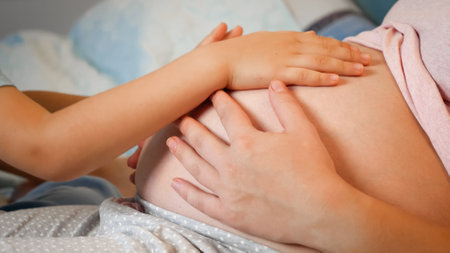 Closeup of little toddler boy stroking and touching big belly of pregnant mother lying in bed