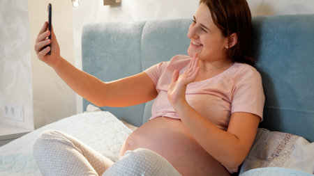 Happy smiling pregnant woman lying in bed and talking to a friend on video conference. Pregnant girl...