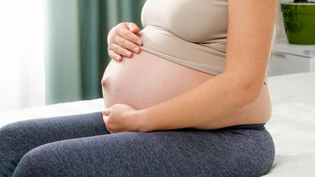 Beautiful pregnant woman sitting on bed side and stroking her bog belly against big window. Concept of healthy lifestyle, healthcare and sports during pregnancyの写真素材