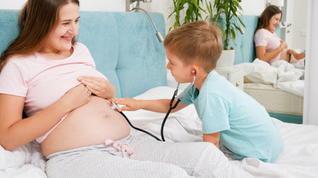 Little toddler boy pretending to be a doctor using stethoscope to listen heartbet of his unborn brother in big belly of pregnant mother. Concept of healthcare and medical examination during pregnancyの写真素材