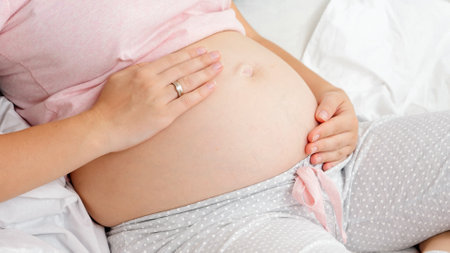 Closeup of young pregnant woman stroking her big belly while sitting on bed. Concept of parenting...