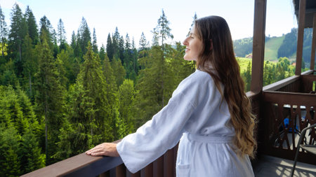 Portrait of smiling woman in bathrobe enjoying view on mountains from her hotel room. Concept of tourism and vacation in mountains at summerの写真素材