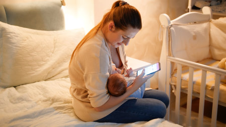 Panning shot of mother using smartphone giving breast to her hungry baby son on bed at night....