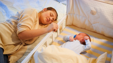 Young tired mother fallen asleep while rocking her baby son in crib at late night. Motherhood and...
