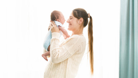 Happy smiling woman with little 1 months old baby boy hugging and embracing at big windw in bedroom. Concept of family happiness and loving parents with little childrenの写真素材