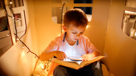 Little boy in toy house with light garlands reading book at night. Concept of child education and reading in dark roomの写真素材