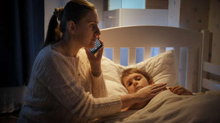 Young worried mother writing to doctor on smartphone next to sick little son lying in bed. Concept of children illness, disease and parent care.の写真素材