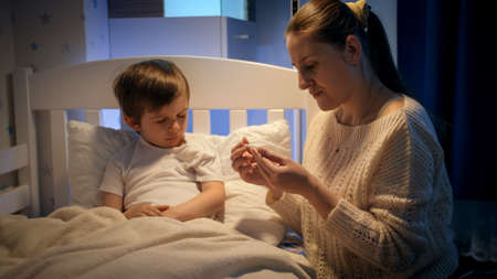 Young caring mother measuring temperature and kissing her sick little boy lying in bed. Concept of...