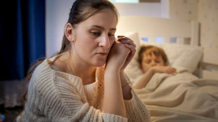 Young mother sitting next to bed of her sleeping son suffering from depression and bad thoughts. Concept of lonely mother, family problems and maternal depressionの写真素材