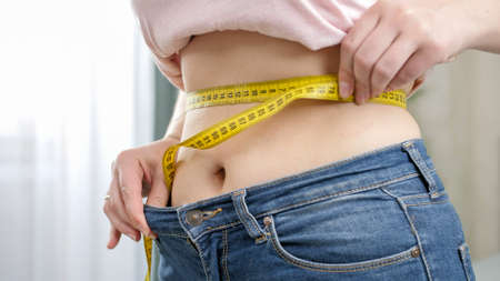 Closeup of young woman with slim belly measuring her waist at mirror in bedroom. Concept of dieting, loosing weight and healthy lifestyle.の写真素材