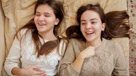 Two happy smiling teenage girls relaxing on bed and chatting. Teens having fun and good time togetherの写真素材