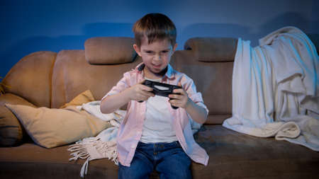 Funny little boy holding game controller while playing video games on TV console at night