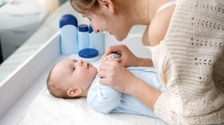 Smiling young mother smiling and talking to her little baby boy lying on dressing table. Concept of hygiene, baby care and healthcareの写真素材