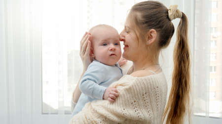 Portrait of beautiful smiling woman hugging and stroking her little baby son at big window. Concept of family happiness and child development.の写真素材