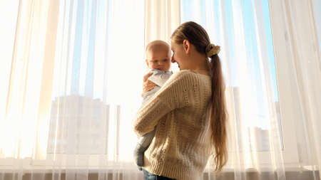 Portrait of beautiful mother holding and kissing her little baby son at big window. Concept of...