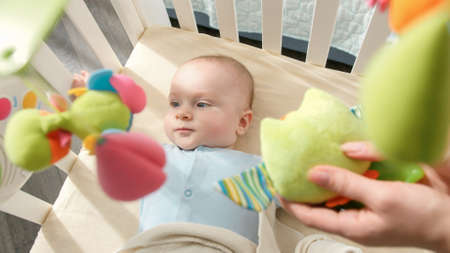 Young mother showing colorful plush toys to her little baby son lying in crib. Concept of baby...