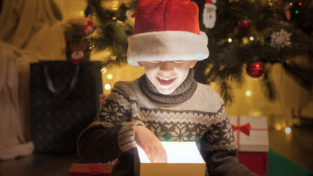Happy smiling boy taking Chrsitmas present out of glowing magical gift box under Christmas treeの写真素材