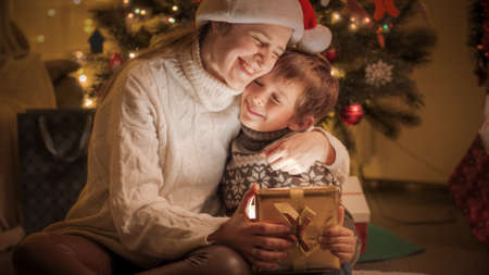 Happy smiling mother and son hugging after getting Christmas presents under Christmas tree at homeの写真素材