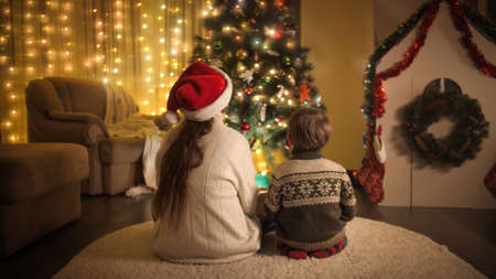 Rear view of happy family sitting on floor and looking on colorful Christmas tree lights. Families and children celebrating winter holidays.の写真素材