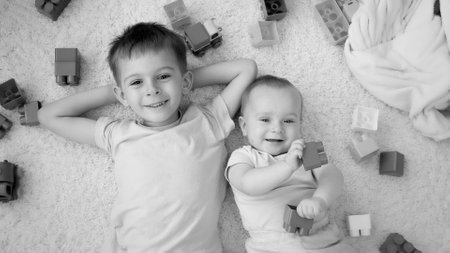 Black and white image of cheerful smiling boy with baby brother surrounded with toys lying on carpet in playroom. Concept of children development, education and creativity at homeの写真素材