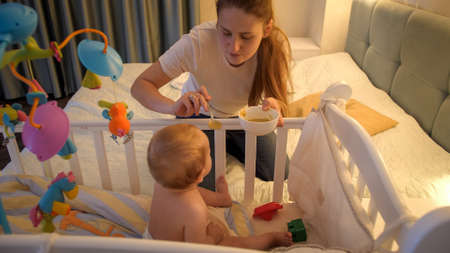 Young caring mother feeding her baby son with soup in baby crib at night. Concept of healthy...