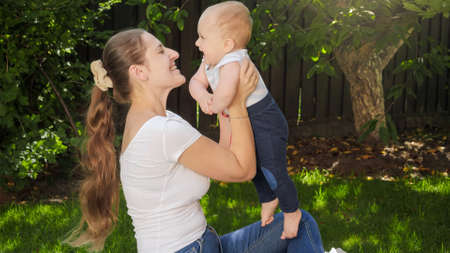 Happy cheerful mother holding and lifting up her little baby son in backyard garden. Parenting, family, children development, and fun outdoors on nature.の写真素材