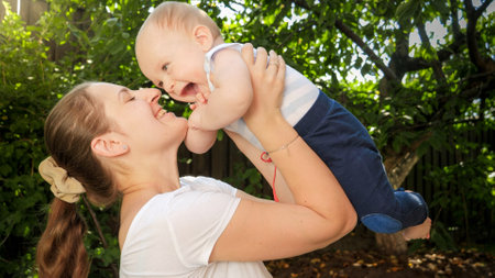 Portrait of smiling woman lifting up her laughing baby boy under trees at parkの写真素材