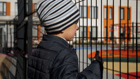 Little upset boy looking through metal fence on modern school and playground. Concept of poverty,...