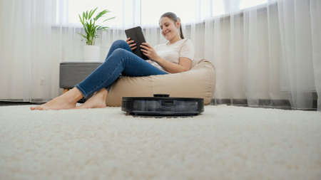 Happy smiling woman relaxing in chair and browsing internet on tablet computer while robot vacuum cleaner doing housework and cleanupの写真素材