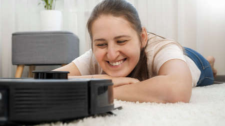 Portrait of happy smiling woman lying on carpet at living room and looking on working robot vacuum cleaner doing houseworkの写真素材