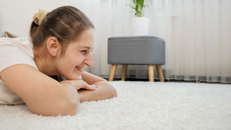 Portrait of happy smiling woman lying on soft white carpet in living room looking to the side. Copy space for your imageの写真素材