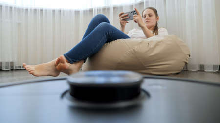 POV shot of robot vacuum cleaner doing house cleanup while young woman relaxing and using...