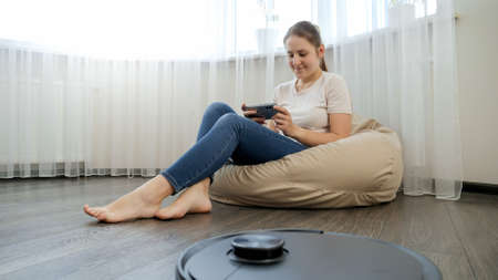 robot vacuum cleaner doing housework while young woman relaxing and resting with smartphone in beanbag chair. Concept of hygiene, household gadgets and robots at modern life.の写真素材
