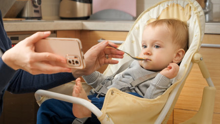 Portrait of little baby boy watching cartoons on smartphone while eating porridge in highchair. Concept of parenting, healthy nutrition and baby feedingの写真素材