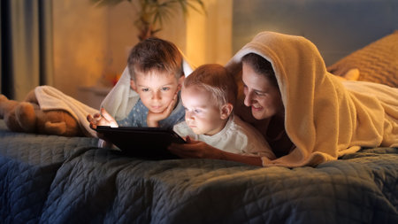 Two boys in pajamas having fun with mother in bed and playing games on tablet computer. Family having time together, parenting, happy childhood and entertainmentの写真素材