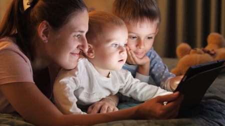 Two smiling brothers in pajamas playing on tablet computer with mother before going to sleep. Family having time together, parenting, happy childhood and entertainmentの写真素材