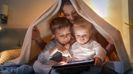Smiling family hiding under blanket at night and using tablet computer. Family having time together, parenting, happy childhood and entertainmentの写真素材