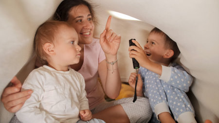 Two cheerful boys having fun with young mother under blanket and playing with flashlight. Family having time together, parenting, happy childhood and entertainmentの写真素材
