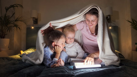 Two boys and mother in pajamas reading bedtime story book in bed at night with flashlights. Family having time together, parenting, happy childhood and entertainmentの写真素材