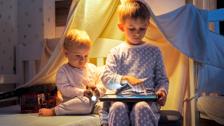 Two cute boys in pajamas playing games on tablet computer in bed at night. Children with gadgets, education, kids development.の写真素材
