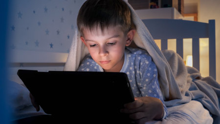 Portrait of little boy lying under blanket and using tablet computer. Children education,...