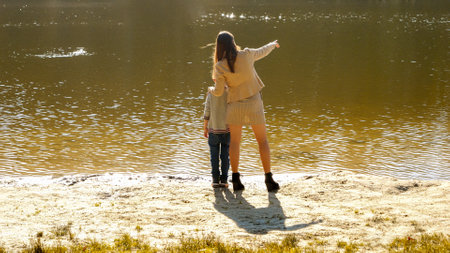 Young mother relaxing with her son at river and showing him fish swimming in water. Family outdoors, happy parenting and childhood, autumn landscape, kids and parents relaxing outsideの写真素材