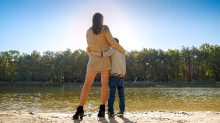 Young mother hugging and kissing her son standing at the lake in the forest. Family outdoors, happy parenting and childhood, autumn landscape, kids and parents relaxing outsideの写真素材