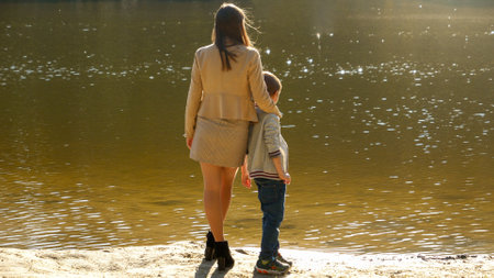 Little boy with young mother standing at the lake and looking on sunset. Family outdoors, happy parenting and childhood, autumn landscape, kids and parents relaxing outsideの写真素材