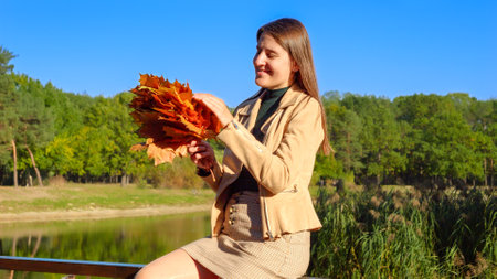 Happy smiling woman holding bunch of autumn tree leaves while relaxing at river. Beauty in nature, fall in the city, happy women.の写真素材
