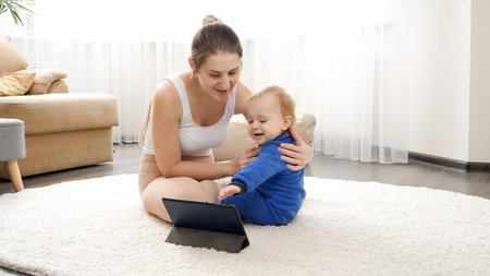 Smiling young mother doing baby massage and body stretching to her little son while watching video...