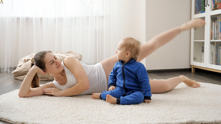 Beautiful smiling woman lying on carpet with baby son and doing fitness exercises. Family healthcare, active lifestyle, parenting and child development.の写真素材