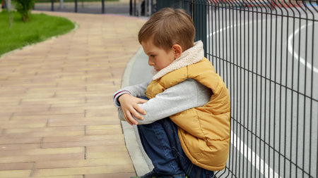 Unhappy lonely boy leaning on metal fence at school playground and sitting down. Child depression, problems with bullying, victim in school, emigration, criminal and poverty.の写真素材